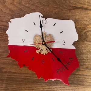 May include: A wooden wall clock shaped like the map of Poland, painted in the colors of the Polish flag: white and red. The clock features black numerals, black hands, and a gold-colored Polish eagle emblem. The word "POLSKA" is printed on the red section.