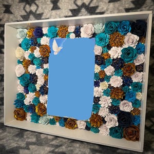 May include: A white rectangular shadow box filled with paper flowers in shades of teal, white, navy, and gold. A blue rectangle is in the center, with a white dove graphic in the upper left corner.