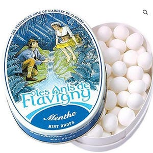 Puede incluir: Una lata ovalada de caramelos de menta Les Anis de Flavigny, con un diseño azul y blanco que presenta una escena pastoral. La lata está abierta, revelando un recipiente lleno de mentas blancas y redondas. El texto "Menthe MINT DROPS" es visible.