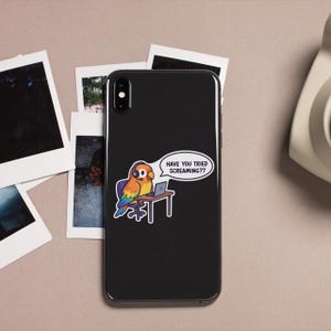 May include: Black smartphone with a sticker featuring a cartoon parrot wearing a headset and working on a laptop. The parrot is sitting at a desk with the text "HAVE YOU TRIED SCREAMING??" in a speech bubble. Several polaroid photos are in the background.