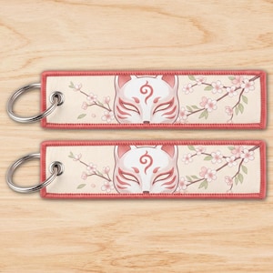 May include: Two rectangular fabric keychains with a cream background, featuring a stylized white and pink fox mask graphic centered between delicate pink cherry blossom branches. Each keychain has a silver metal ring and a pink stitched border.