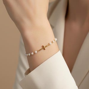 May include: A gold-colored bracelet featuring a small cross pendant and white pearls. The bracelet is worn on a wrist, with the cross positioned centrally. The pearls are evenly spaced along the bracelet's chain, creating a delicate and elegant design.