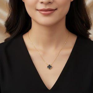 May include: A gold-colored necklace with a four-leaf clover pendant. The pendant is black and sits at the center of the chest. The necklace is worn against a black top.