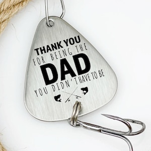 May include: A silver-colored fishing lure with the text "THANK YOU FOR BEING THE DAD YOU DIDN'T HAVE TO BE." The lure is attached to a metal swivel and two hooks. A length of tan rope is coiled around the lure.