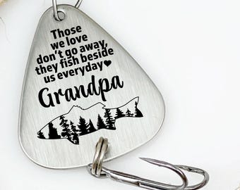 May include: A silver-colored fishing lure with a hook and swivel, featuring the text "Those we love don't go away, they fish beside us everyday" and the word "Grandpa" with a fish and forest graphic. The lure is set against a backdrop of a thick, tan rope.