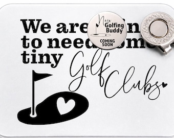 Pode incluir: Uma caixa de presente retangular branca com tema de golfe com o texto "We are going to need some tiny Golf Clubs". Um marcador de bola prateado e um marcador de bola de golfe com o texto "New Golfing Buddy Coming Soon" estão em cima.