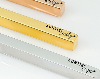 May include: Three personalized bar necklaces in rose gold, gold, and silver. Each necklace features the word "AUNTIE" followed by a name, such as "Kailyn", "Emily", and "Elyn". The necklaces are displayed on a white surface.