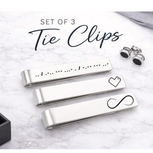May include: Set of three silver tie clips with different designs. One clip has Morse code, another a pixelated heart, and the last an infinity symbol. The words "Tie Clips" are above the clips. Also included are three black and silver cufflinks.