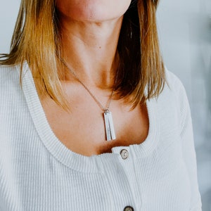 May include: A silver necklace with two rectangular pendants hanging from a delicate chain. The necklace is worn by a person with long brown hair and a white shirt.