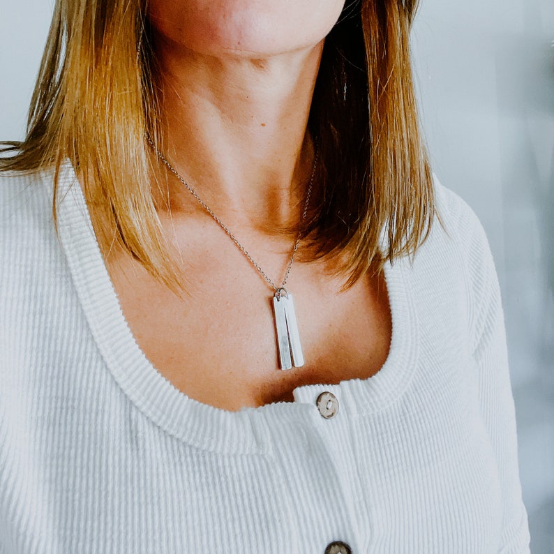 May include: A silver necklace with two rectangular pendants hanging from a delicate chain. The necklace is worn by a person with long brown hair and a white shirt.