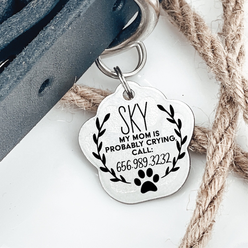 Silver Pawprint shaped pet tag with "SKY MY MOM IS PROABLY CRYING, CALL: 656.989.3232" engraved on to it. Hole in top of pet tag attaches to two jump rings that attach to a dog collar.