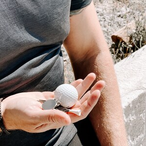 May include: A white golf ball, tee, and keychains are held in an open hand. The golf ball has a dimpled surface. The tee is white. The keychains have a silver finish. The person is wearing a gray t-shirt and a black bracelet.