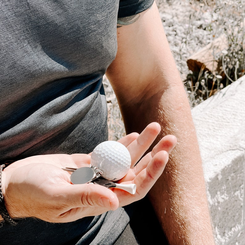 May include: A white golf ball, tee, and keychains are held in an open hand. The golf ball has a dimpled surface. The tee is white. The keychains have a silver finish. The person is wearing a gray t-shirt and a black bracelet.