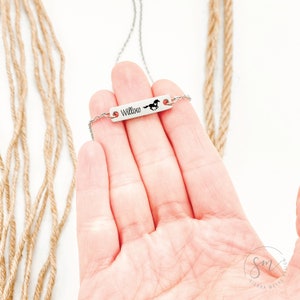 K&ouml;nnte beinhalten: Eine silberne Halskette mit einem kleinen rechteckigen Anh&auml;nger, auf dem "Willow" steht und eine Pferdesilhouette abgebildet ist. Die Halskette liegt auf einer Hand vor einem Hintergrund aus braunem Seil.