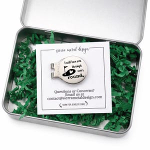 May include: A silver golf ball marker with the text "I will love you through every round" inside a metal tin filled with green paper shreds. The marker has a golf flag and heart design. A white card with the text "sierra metal design" is also in the tin.