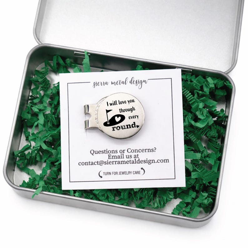 May include: A silver golf ball marker with the text "I will love you through every round" inside a metal tin filled with green paper shreds. The marker has a golf flag and heart design. A white card with the text "sierra metal design" is also in the tin.