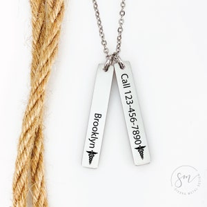May include: A silver necklace with two vertical bars. The left bar is engraved with the name "Brooklyn" and a medical symbol. The right bar is engraved with the text "Call 123-456-7890" and a medical symbol.