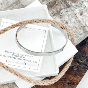 May include: A silver bangle bracelet in a white box with a brown rope wrapped around it. The box has a card inside with the text "Questions or Concerns? Email us at contact@sierrametaldesign.com (TURN FOR JEWELRY CARE".