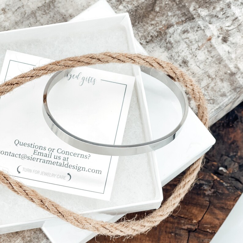 May include: A silver bangle bracelet in a white box with a brown rope wrapped around it. The box has a card inside with the text "Questions or Concerns? Email us at contact@sierrametaldesign.com (TURN FOR JEWELRY CARE".