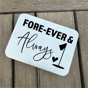 May include: A rectangular, light-colored metal sign with rounded corners. The sign features the words "FORE-EVER & Always 1" in black text and a black golf flag and heart symbol. The sign is placed on a wooden surface.