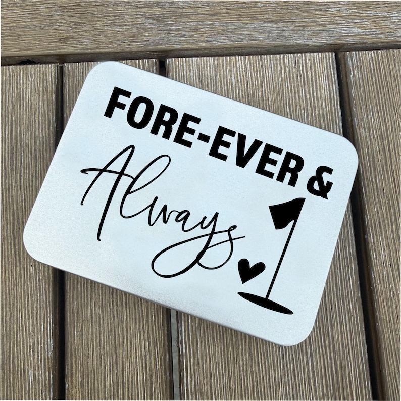 May include: A rectangular, light-colored metal sign with rounded corners. The sign features the words "FORE-EVER & Always 1" in black text and a black golf flag and heart symbol. The sign is placed on a wooden surface.
