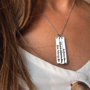 May include: A silver necklace with three vertical bar pendants. Each pendant has a medical symbol and text that reads "Asthma", "Wheat Allergy", and "Bee Allergy".