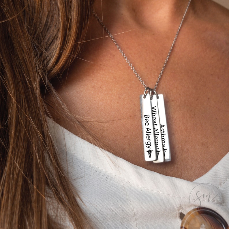 May include: A silver necklace with three vertical bar pendants. Each pendant has a medical symbol and text that reads "Asthma", "Wheat Allergy", and "Bee Allergy".