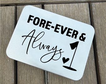May include: A rectangular, light-colored metal sign with rounded corners. The sign features the words "FORE-EVER & Always 1" in black, stylized lettering. A black heart and a golf flag graphic are also present.