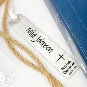 May include: A silver bookmark with the name "Mia Johnson" engraved on it, along with a cross and the text "First Holy Communion April 7th, 2025".
