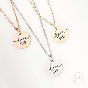 May include: Three silver necklaces with round pendants that say "love" with initials and a small heart. The necklaces are on a white background.