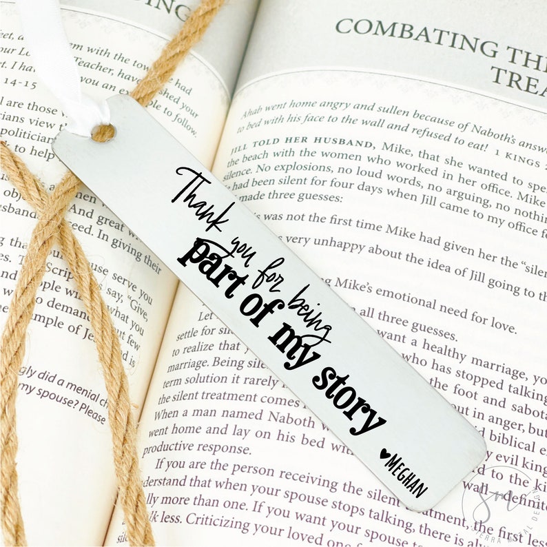 May include: A white metal bookmark with black text that reads "Thank you for being part of my story". The bookmark is laying on a book with text about a husband and wife. The bookmark has a small heart with the name "Meghan" written below it.