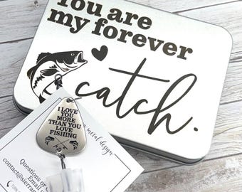 May include: A silver metal box with the words "You are my forever catch" in black script. A fish graphic and heart are also on the box. A fishing-themed badge reel with the text "I love you more than you love fishing" is in front of the box.