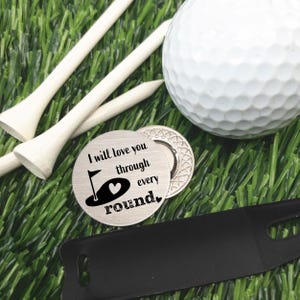 May include: A golf-themed gift set on artificial turf. Includes a white golf ball, wooden tees, a divot repair tool, and a silver golf ball marker with the text "I will love you through every round."