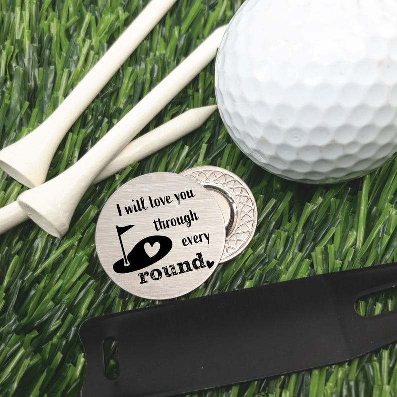 May include: A golf-themed gift set on artificial turf. Includes a white golf ball, wooden tees, a divot repair tool, and a silver golf ball marker with the text "I will love you through every round."
