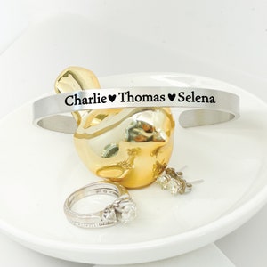 May include: Silver bangle bracelet inscribed with "Charlie &hearts; Thomas &hearts; Selena" on a white surface.