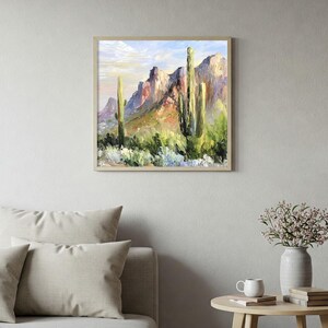 May include: Framed landscape painting depicting a desert scene with towering cacti and rugged mountains. The artwork features a palette of greens, browns, and blues, with a light-colored frame. The painting is displayed above a sofa and side table.