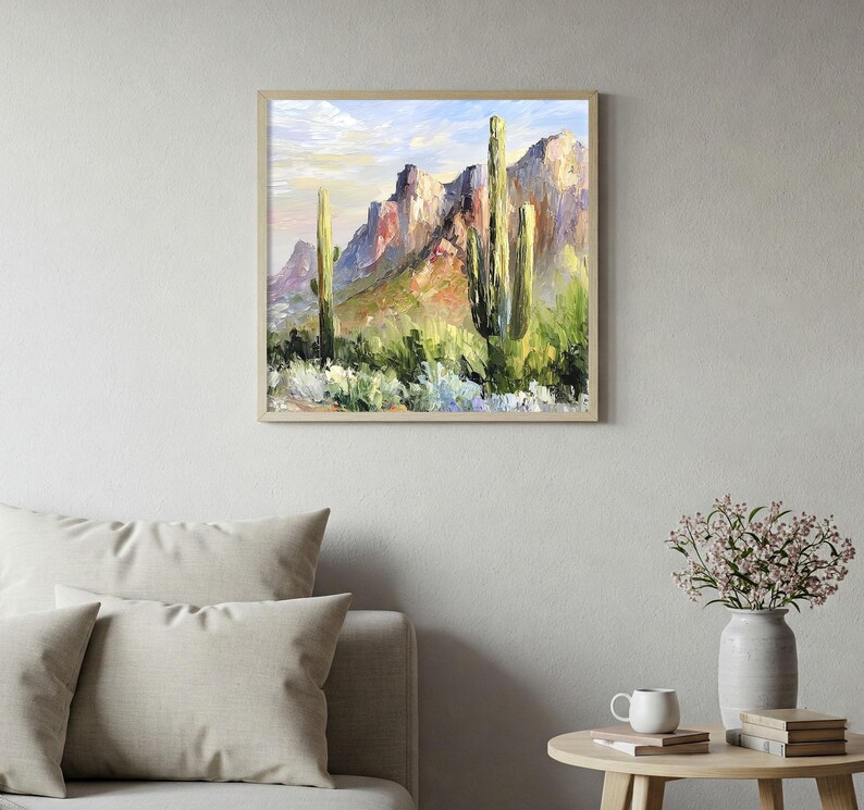 May include: Framed landscape painting depicting a desert scene with towering cacti and rugged mountains. The artwork features a palette of greens, browns, and blues, with a light-colored frame. The painting is displayed above a sofa and side table.