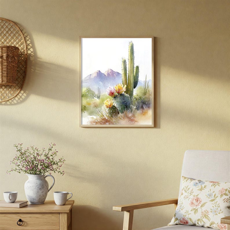 May include: Watercolor painting of a desert landscape featuring cacti with yellow and pink flowers, mountains, and greenery. The artwork is framed in a light wood frame, creating a serene and natural aesthetic. The painting evokes a sense of the American Southwest.