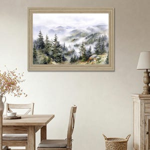 May include: A framed watercolor painting depicts a misty mountain landscape. The artwork features evergreen trees and rolling hills shrouded in fog, with a light-colored wooden frame. The color palette includes greens, blues, and whites.