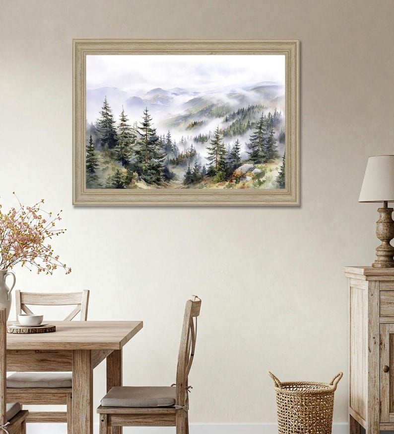 May include: A framed watercolor painting depicts a misty mountain landscape. The artwork features evergreen trees and rolling hills shrouded in fog, with a light-colored wooden frame. The color palette includes greens, blues, and whites.