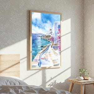 May include: A vertical framed wall art print depicting a coastal Mediterranean scene with white buildings, a stone pathway, vibrant pink bougainvillea flowers, and a bright blue sea under a clear sky with soft white clouds in a light wood frame.