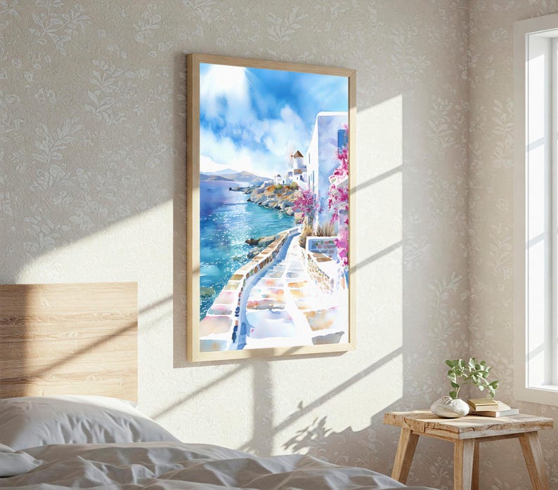May include: A vertical framed wall art print depicting a coastal Mediterranean scene with white buildings, a stone pathway, vibrant pink bougainvillea flowers, and a bright blue sea under a clear sky with soft white clouds in a light wood frame.