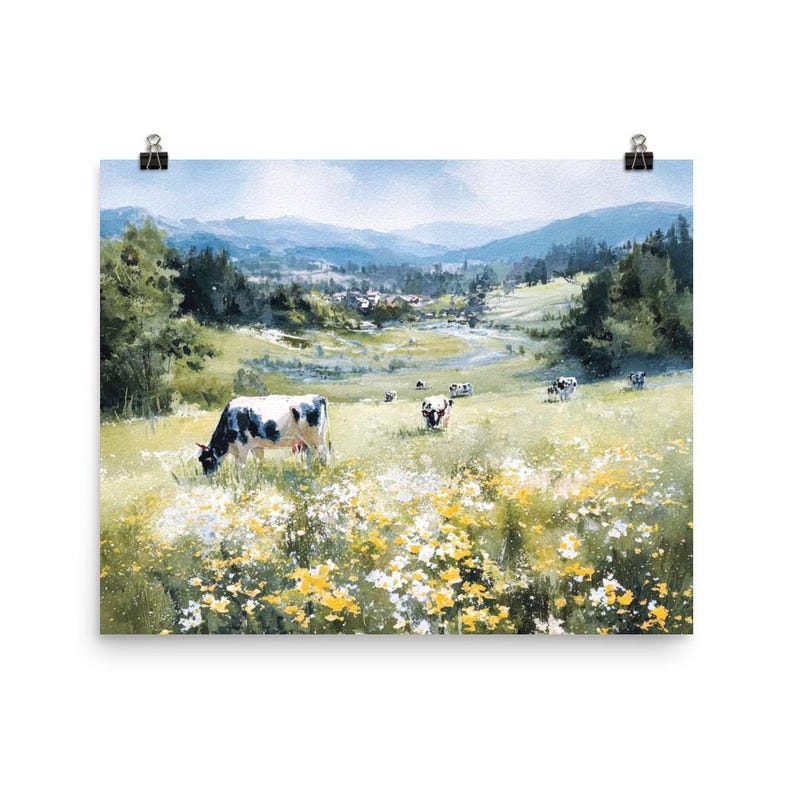 May include: A framed print of a pastoral landscape featuring cows grazing in a field of wildflowers. The artwork depicts a sunny day with rolling hills, trees, and a distant village. The cows are black and white.