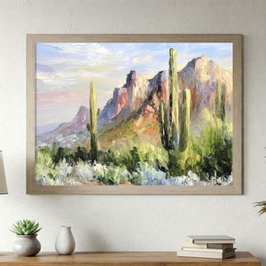 May include: Oil painting of a desert landscape with mountains and cacti. The artwork features a light brown wooden frame. The mountains are painted in shades of purple, red, and yellow, with green cacti in the foreground. The sky has soft pink and blue hues.