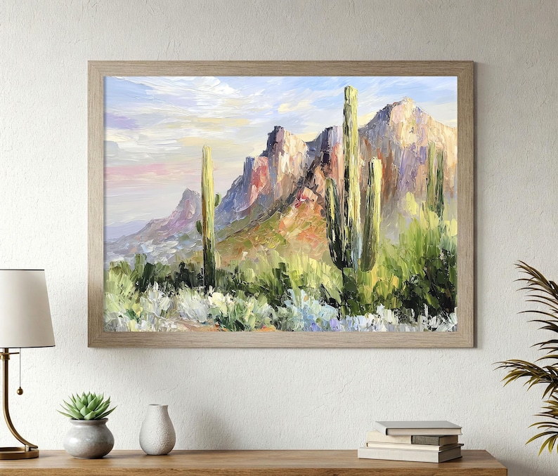 May include: Oil painting of a desert landscape with mountains and cacti. The artwork features a light brown wooden frame. The mountains are painted in shades of purple, red, and yellow, with green cacti in the foreground. The sky has soft pink and blue hues.