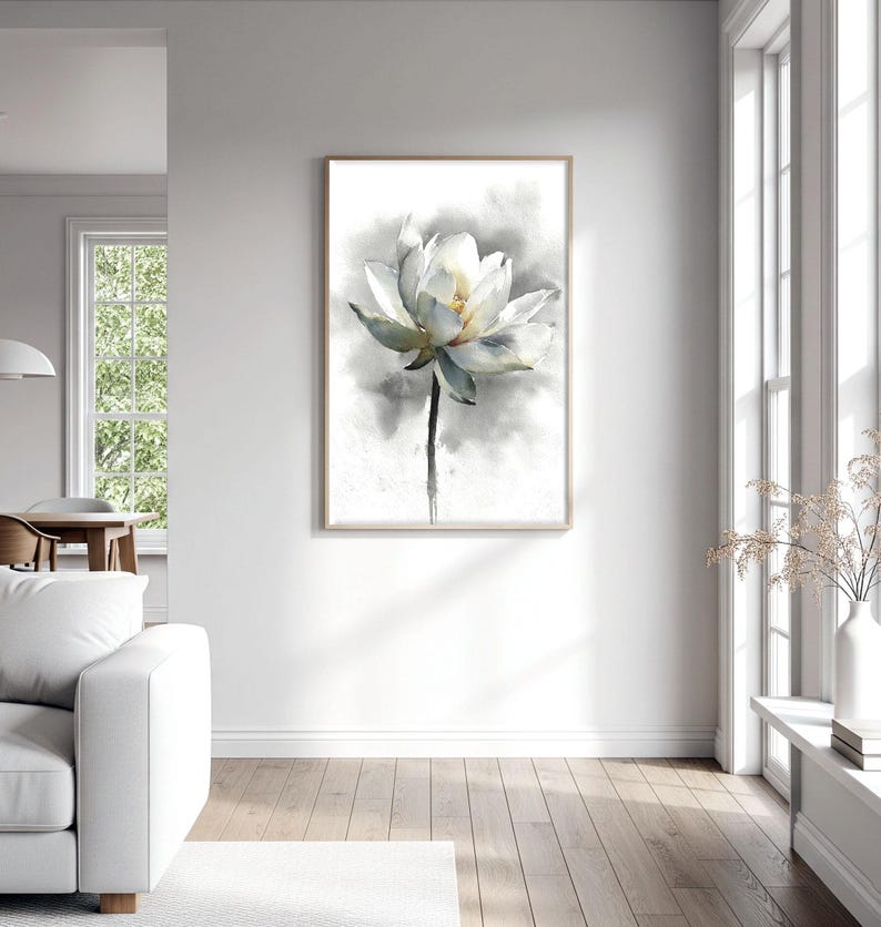 May include: A framed watercolor painting of a white lotus flower with a yellow center and green-blue tinged petals. The flower is set against a soft, gray background. The artwork is displayed in a bright, modern living room.