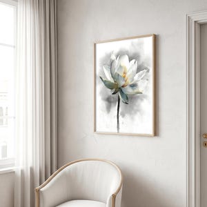 May include: Watercolor painting of a white lotus flower with yellow center and green-blue accents. The flower is set against a soft gray and white background, framed in a light wood frame. The artwork is displayed on a white wall.