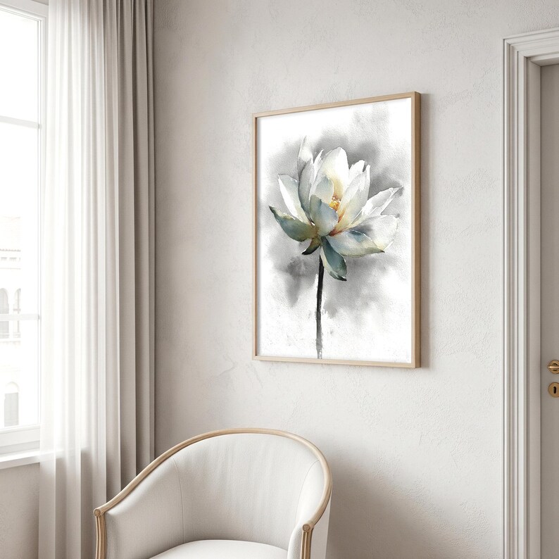 May include: Watercolor painting of a white lotus flower with yellow center and green-blue accents. The flower is set against a soft gray and white background, framed in a light wood frame. The artwork is displayed on a white wall.