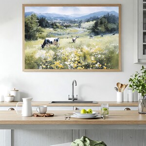 May include: A framed watercolor painting depicts a pastoral scene with cows grazing in a field of wildflowers. The artwork features a light brown wooden frame and is displayed in a kitchen setting. The painting showcases a valley with mountains in the background.
