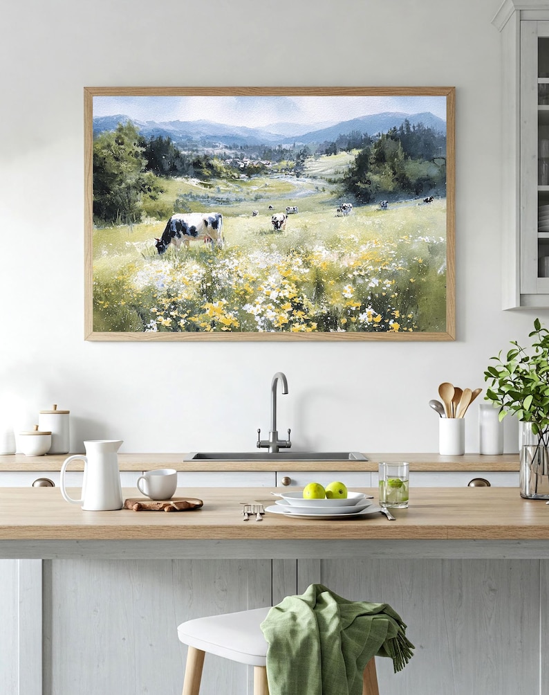 May include: A framed watercolor painting depicts a pastoral scene with cows grazing in a field of wildflowers. The artwork features a light brown wooden frame and is displayed in a kitchen setting. The painting showcases a valley with mountains in the background.